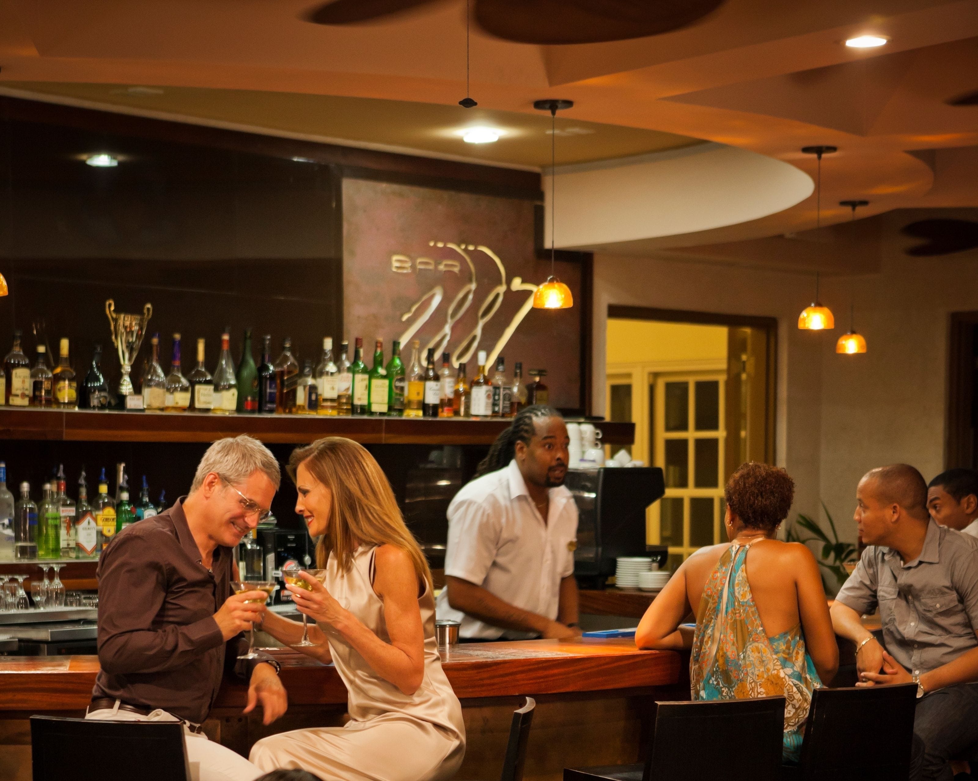 A bar area with a variety of liquor bottles on shelves, a bartender, and patrons seated at tables.