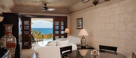 A room with a bed, a dining table, chairs, a chandelier, a vase, and a view of the ocean.