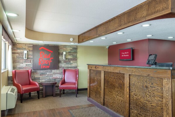A Red Roof Inn reception area with a stone wall backdrop, two leather chairs, a reception desk, and a sign indicating free Wi-Fi in all rooms. - Red Roof Inn Kalamazoo East - Expo Center (Kalamazoo)