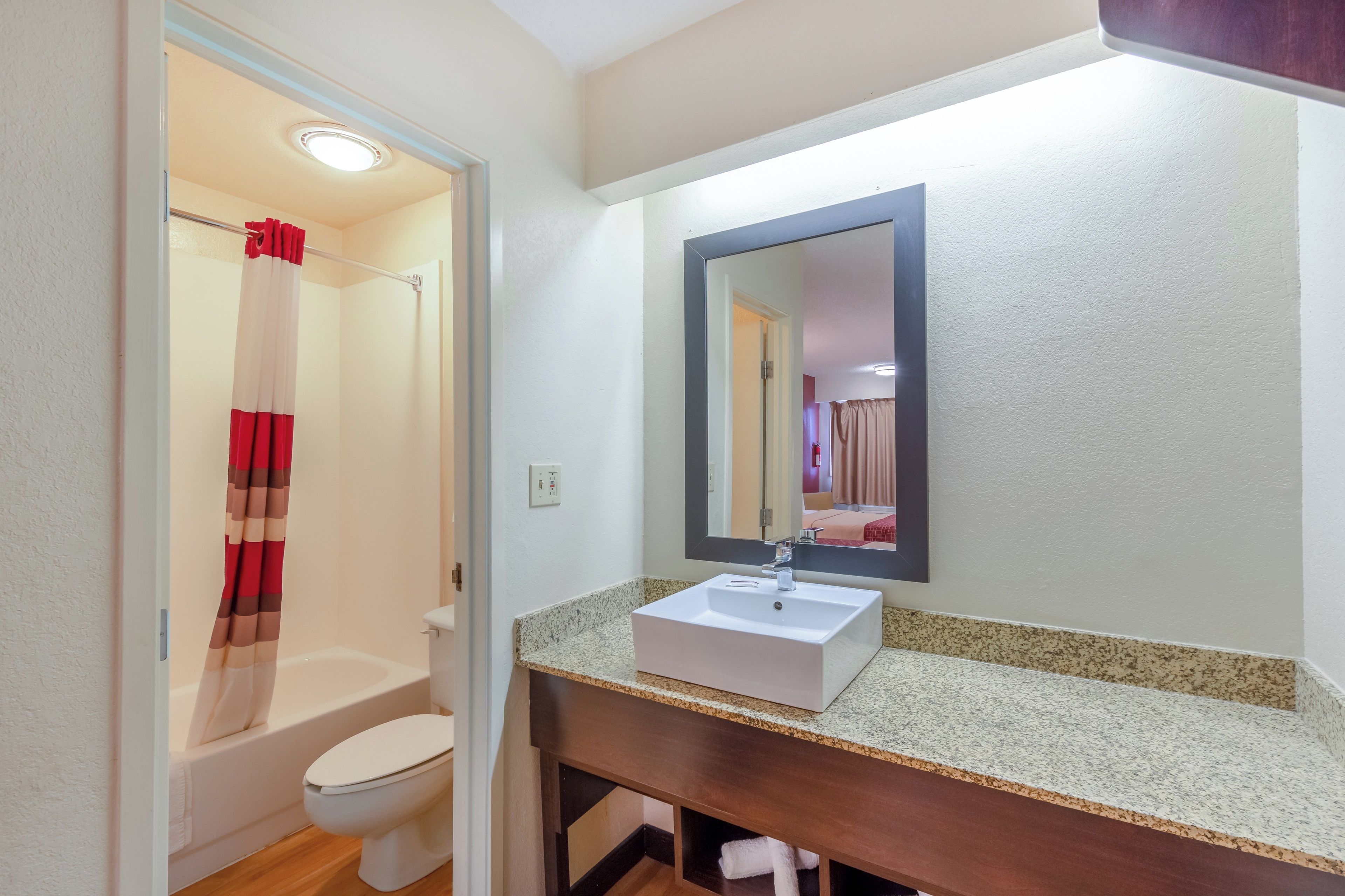 A bathroom with a white toilet, a shower with a red and white curtain, a white sink with a mirror above it, and a granite countertop.