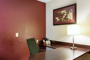 A hotel room with a wooden desk, a green chair, a framed picture on the wall, and a lamp. - Red Roof Inn Kalamazoo East - Expo Center (Kalamazoo)