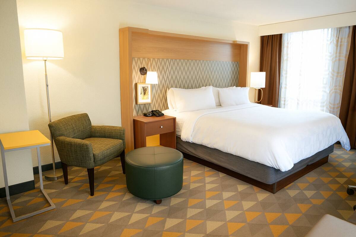 A hotel room with a bed, a chair, a small table, a lamp, and a nightstand.