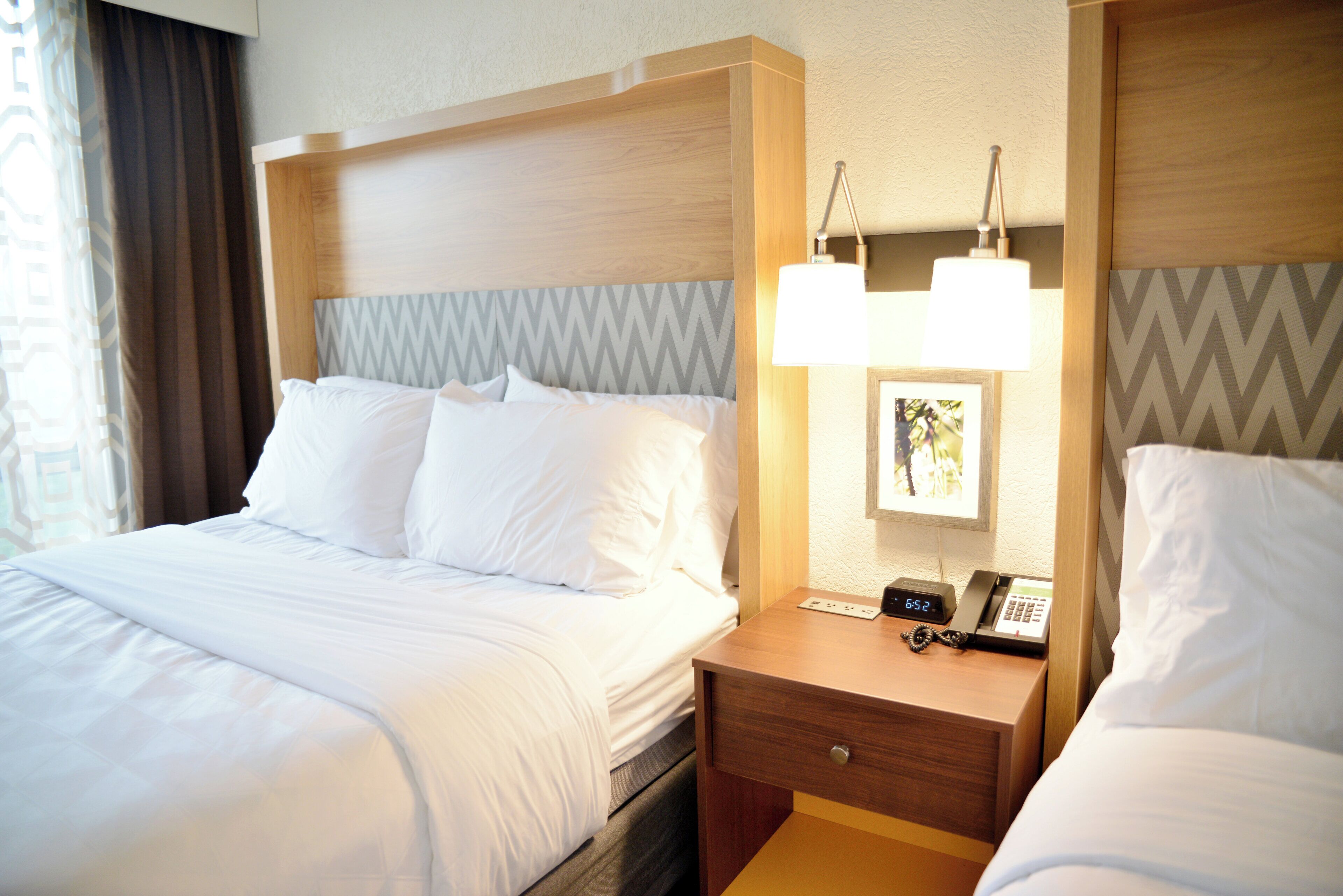Quarto de hotel com cama, mesa de cabeceira, abajur e relógio.