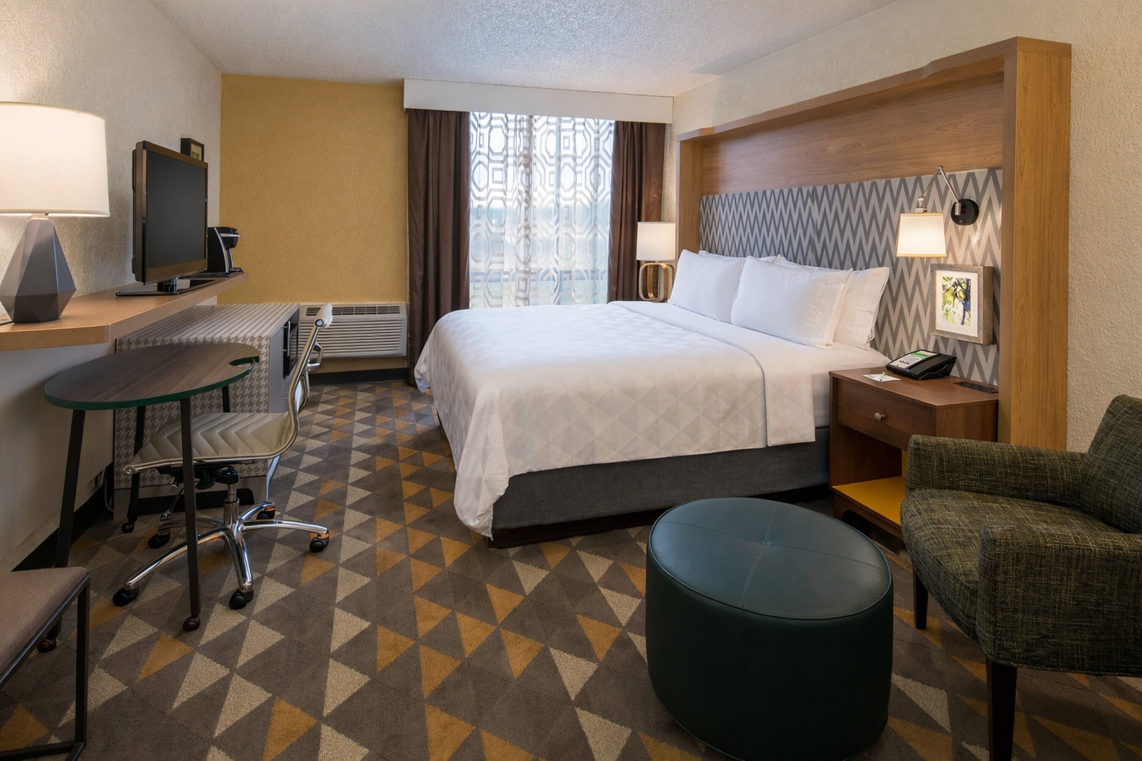A hotel room with a bed, desk, chair, and ottoman.
