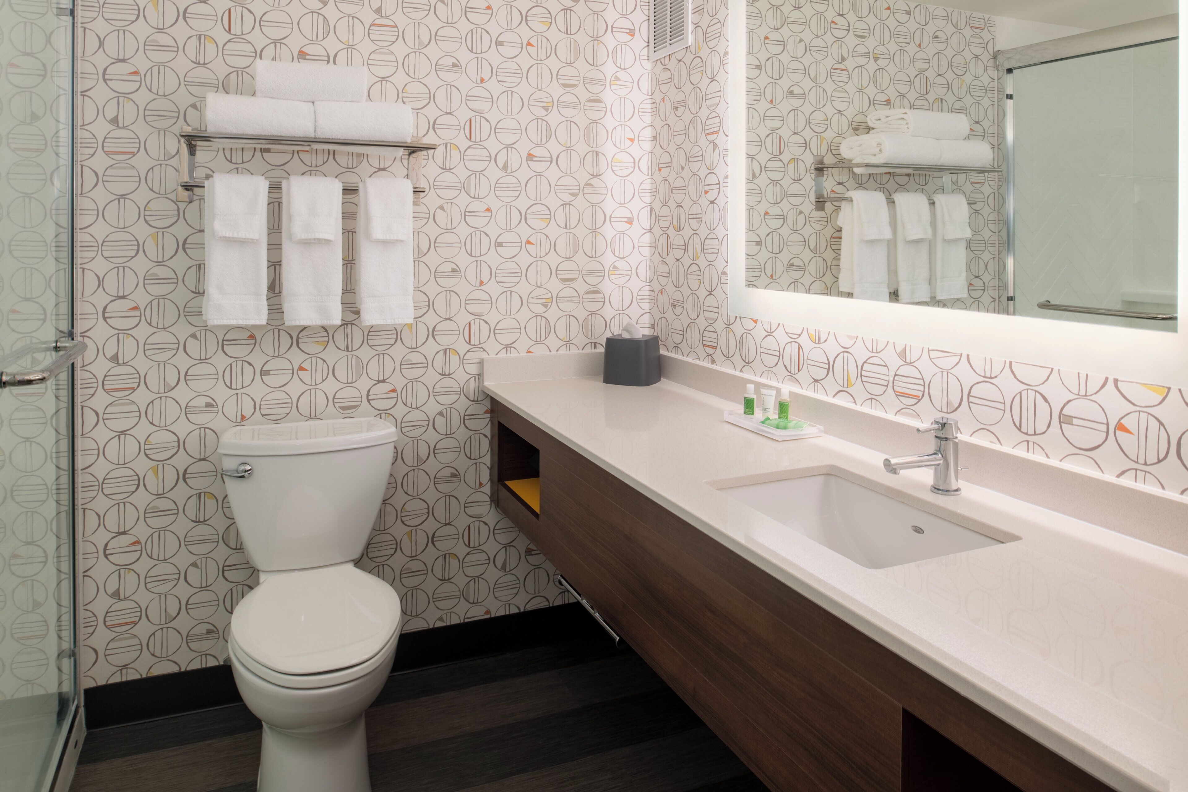 Un baño con inodoro, lavamanos y espejo. Hay toallas en un estante sobre el inodoro.