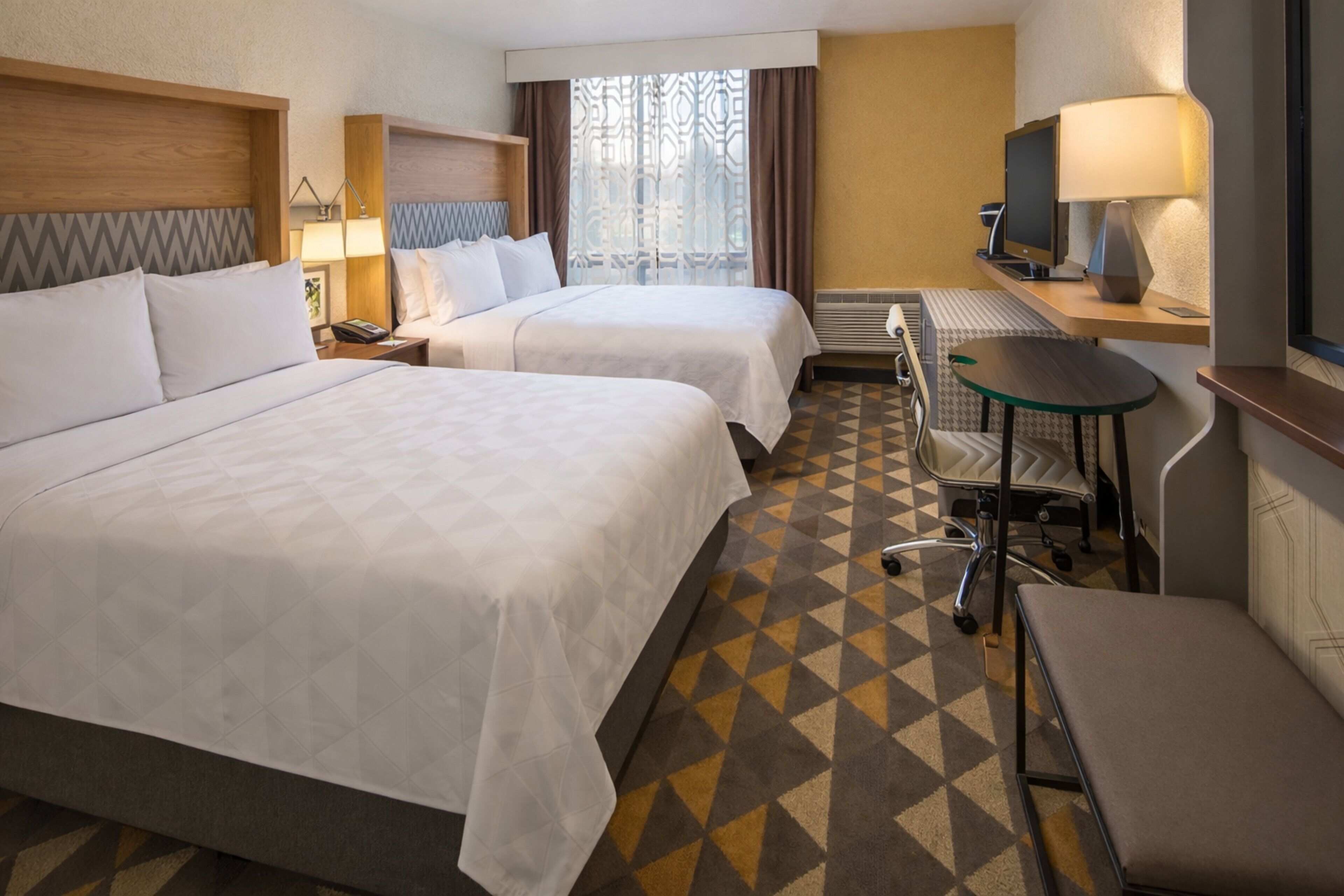 A hotel room with two beds, a desk, a chair, a TV, and a window with curtains.