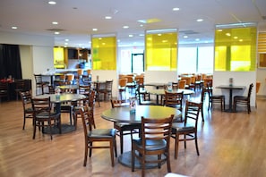 A spacious dining area with multiple round tables and chairs, a kitchen area in the background, and large windows allowing natural light.
