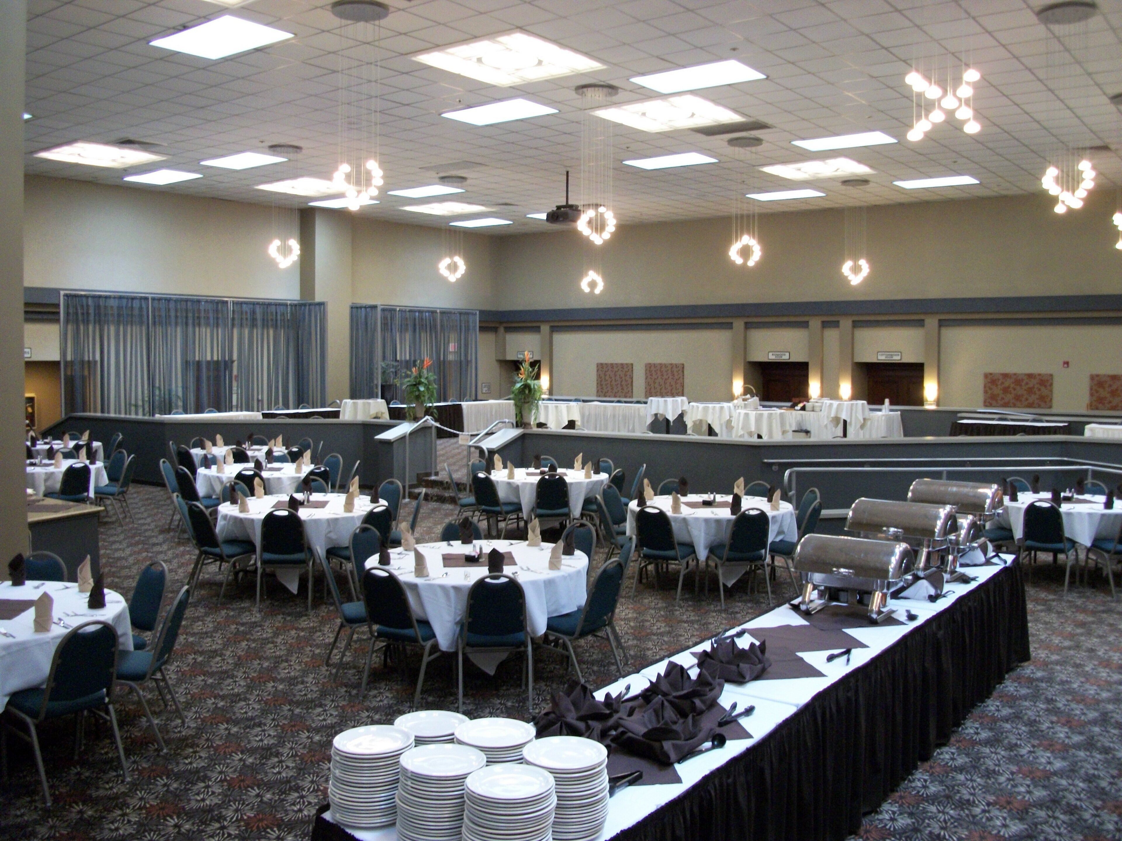 A banquet hall with round tables, chairs, and a long table with stacked plates.
