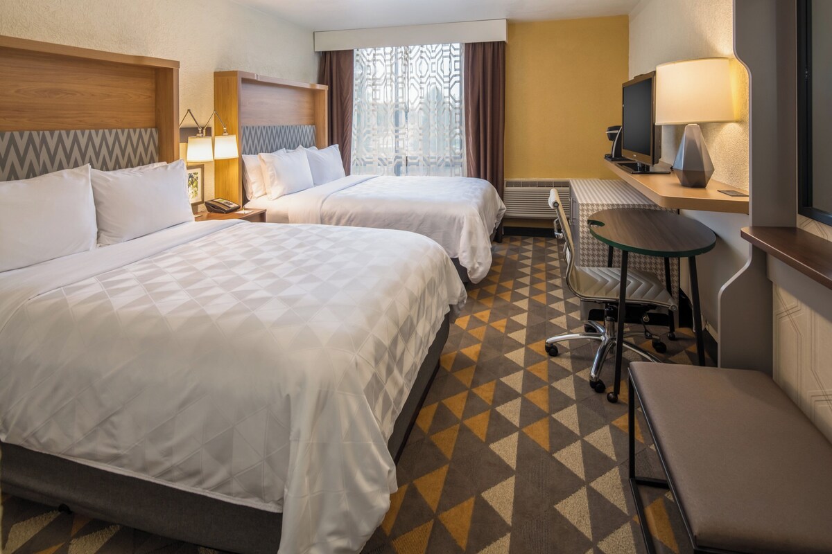 Quarto de hotel com duas camas, uma escrivaninha, uma cadeira, uma televisão e uma janela com cortinas.