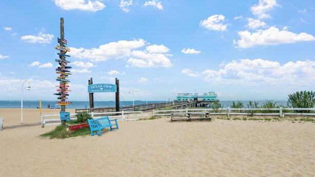A beach with a wooden signpost, a pier, and a blue bench. - Budget Inn Norfolk Va (Norfolk)