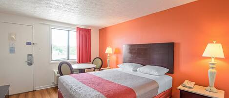 A hotel room with a bed, two bedside lamps, a small table with a chair, a window with curtains, and a door.