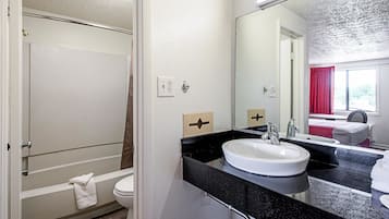 A bathroom with a white sink, black countertop, and a large mirror. There is a towel hanging on the wall and a shower visible in the background.