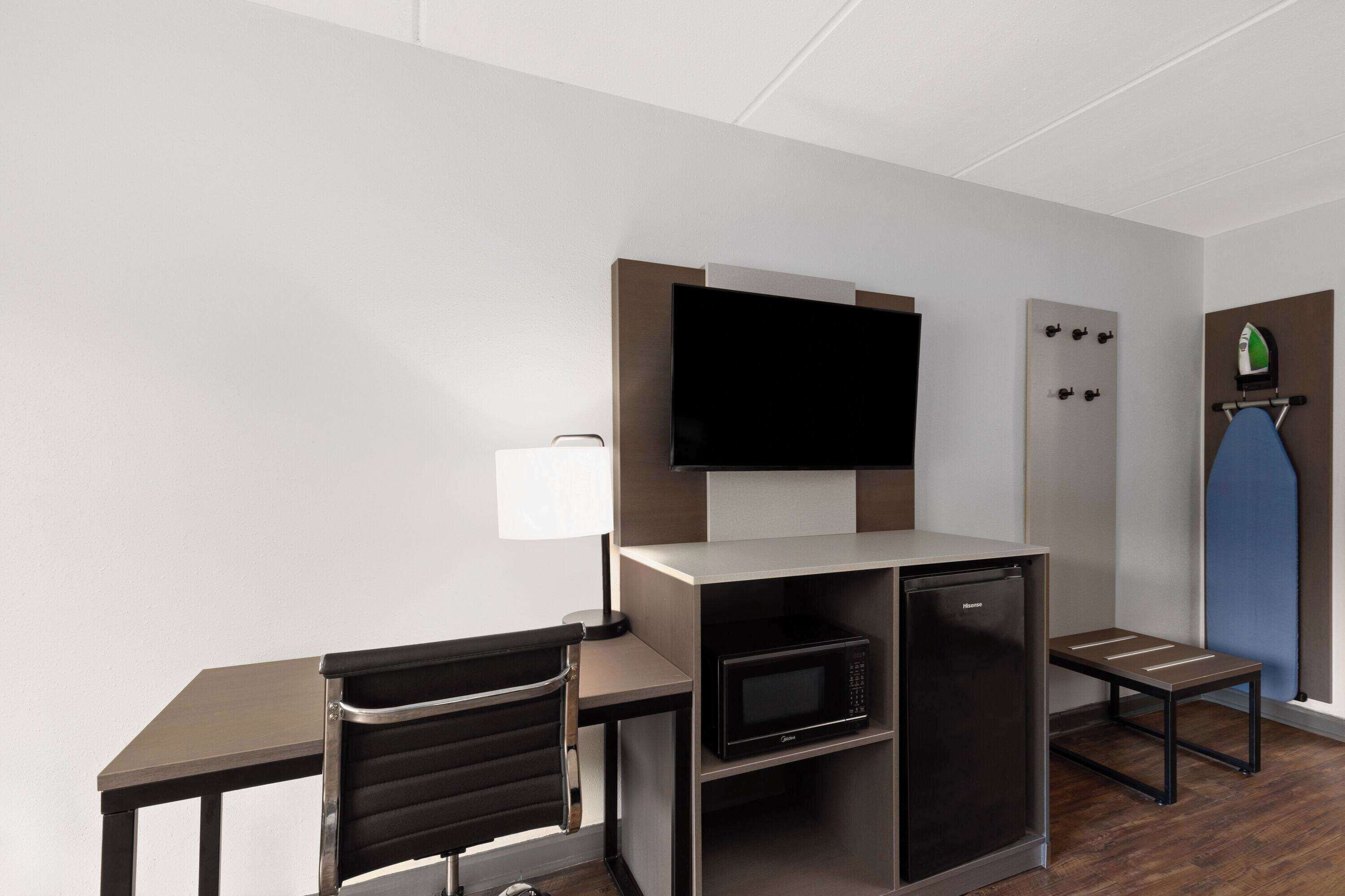 A compact hotel room with a flat-screen TV mounted on the wall, a small microwave, a mini-fridge, a foldable table, a chair, and a small ironing station.