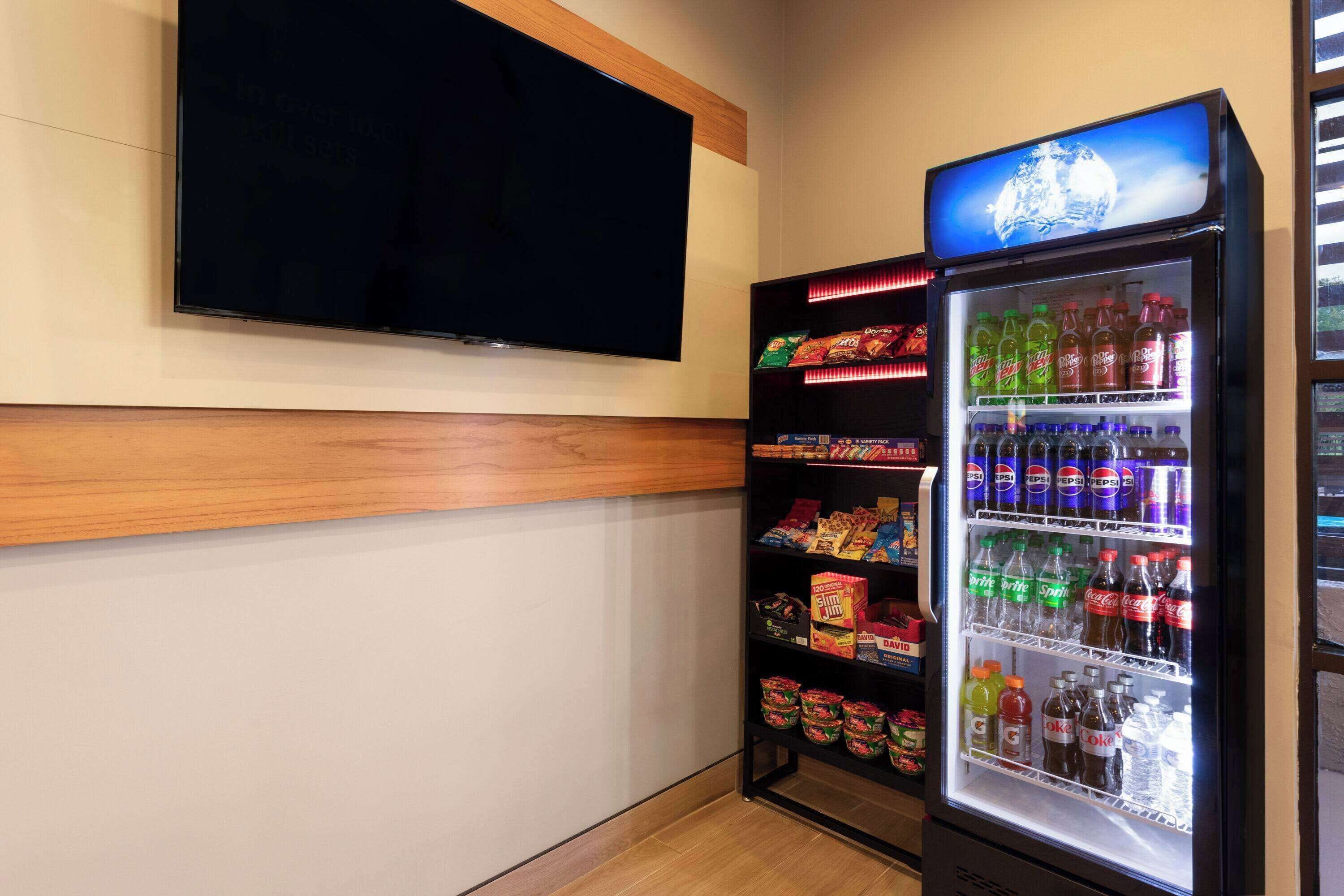 Máquina expendedora llena de bebidas y aperitivos, un televisor montado en la pared, y un diseño de paneles de madera en la pared.