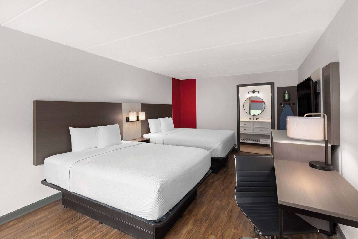 Quarto de hotel com duas camas, uma escrivaninha, uma cadeira, uma TV, um espelho e uma parede de destaque vermelha.