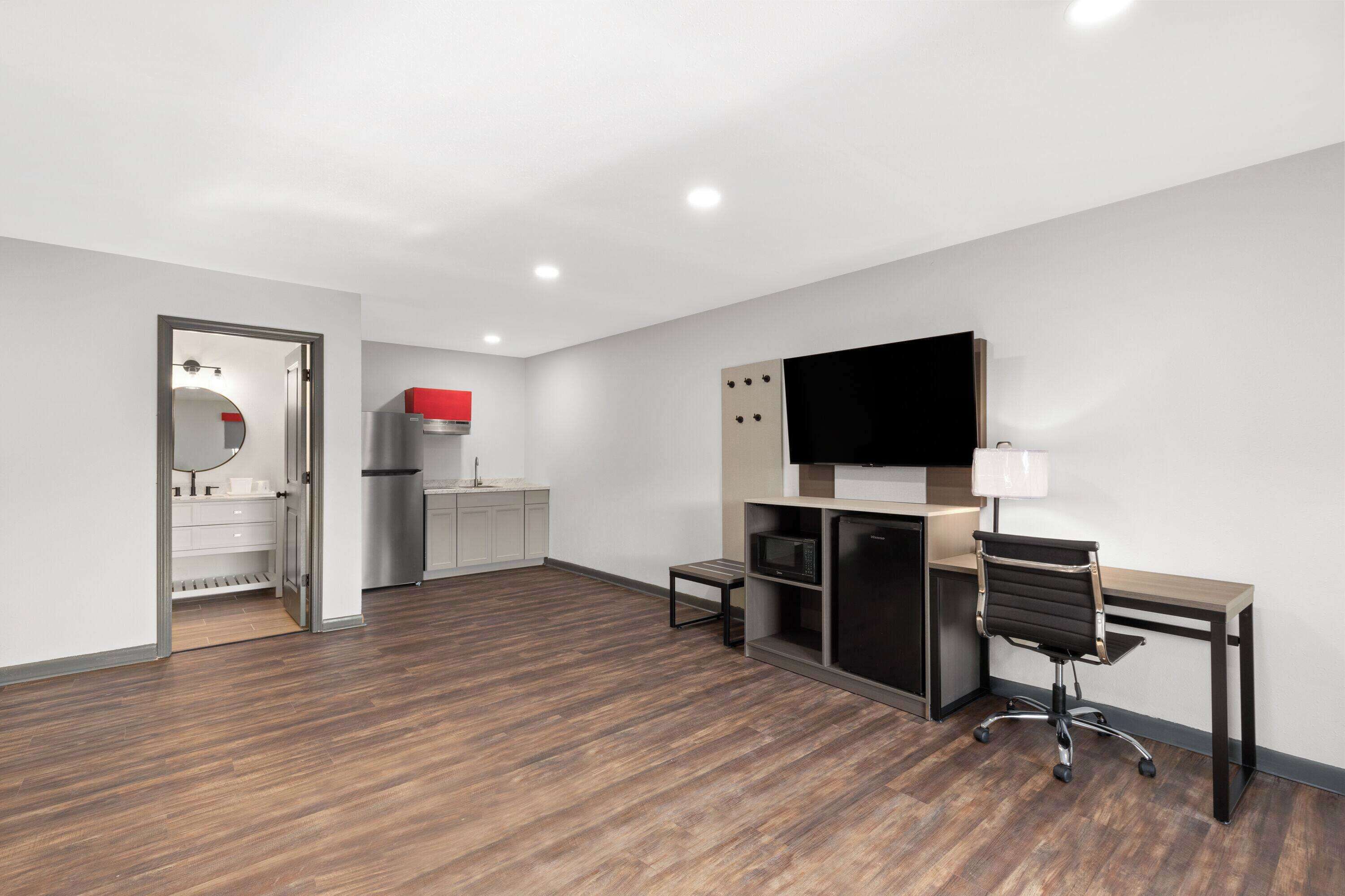A modern hotel room with a wooden floor, a desk with a chair, a flat-screen TV, a microwave, and a refrigerator.