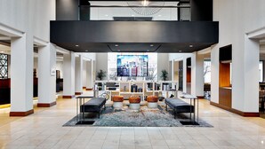 A spacious lobby with a high ceiling, a central seating area with ottomans, a large window with a city view, and a modern chandelier. - Detroit Metro Airport Marriott (Romulus)