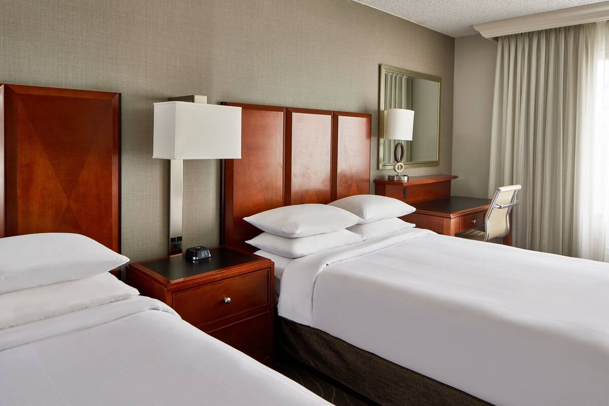 A hotel room with two beds, a wooden headboard, a bedside table with a lamp, a mirror, and a chair.