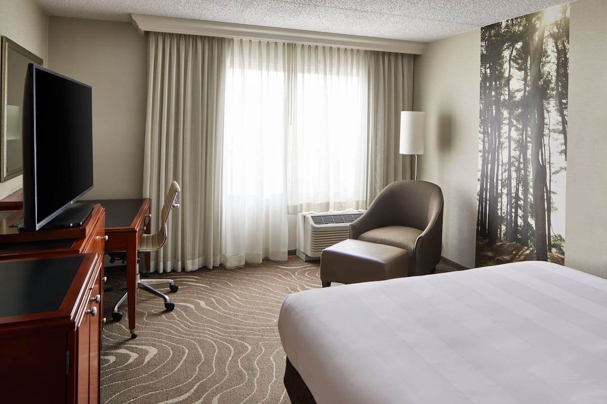 A hotel room with a bed, a desk with a flat-screen TV, a chair, an air conditioning unit, and a large window with curtains.