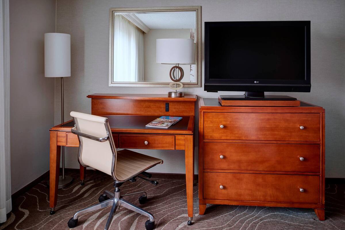 A hotel room with a wooden desk, a chair, a television, a dresser, and a lamp.