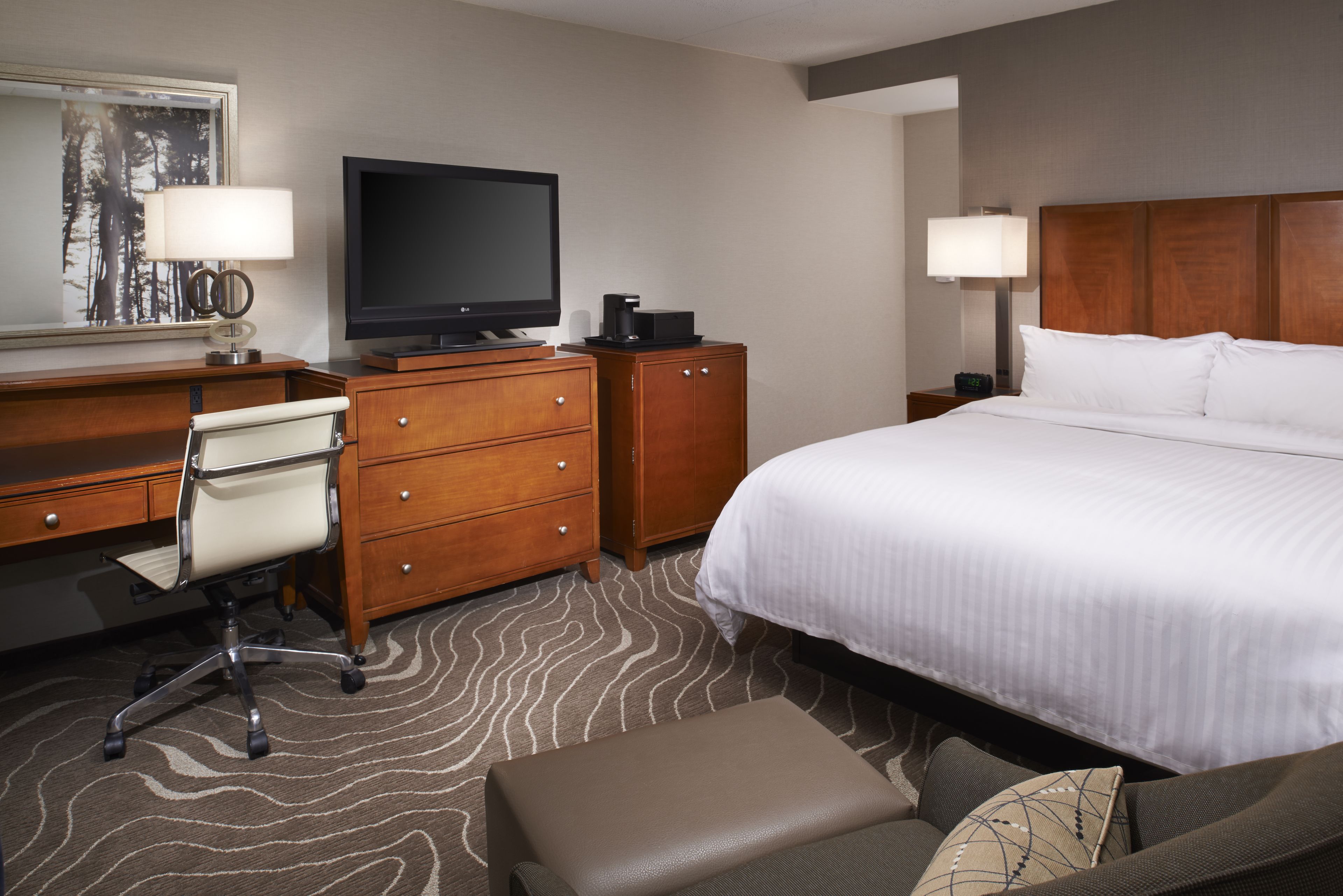Une chambre d’hôtel comprenant un lit, un bureau avec une chaise, une télévision, une commode et une table de chevet avec une lampe.