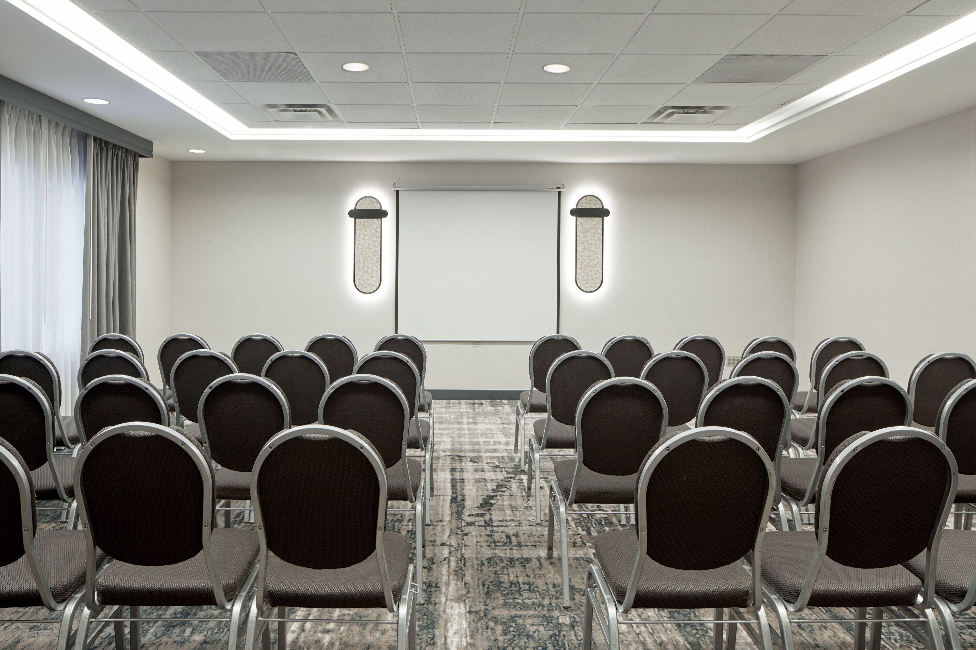 A conference room with rows of chairs facing a projection screen, carpeted floor, and a ceiling with recessed lighting.