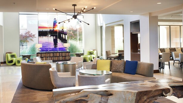 A spacious lobby with a large wooden coffee table, comfortable seating, a bookshelf, and a view of a bridge and sunset through large windows. - Detroit Metro Airport Marriott (Romulus)
