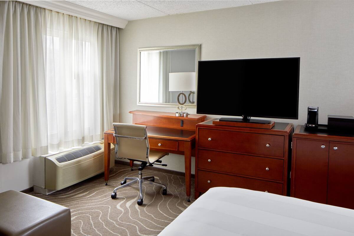 A hotel room with a bed, a desk with a chair, a television, a dresser, and a radiator.