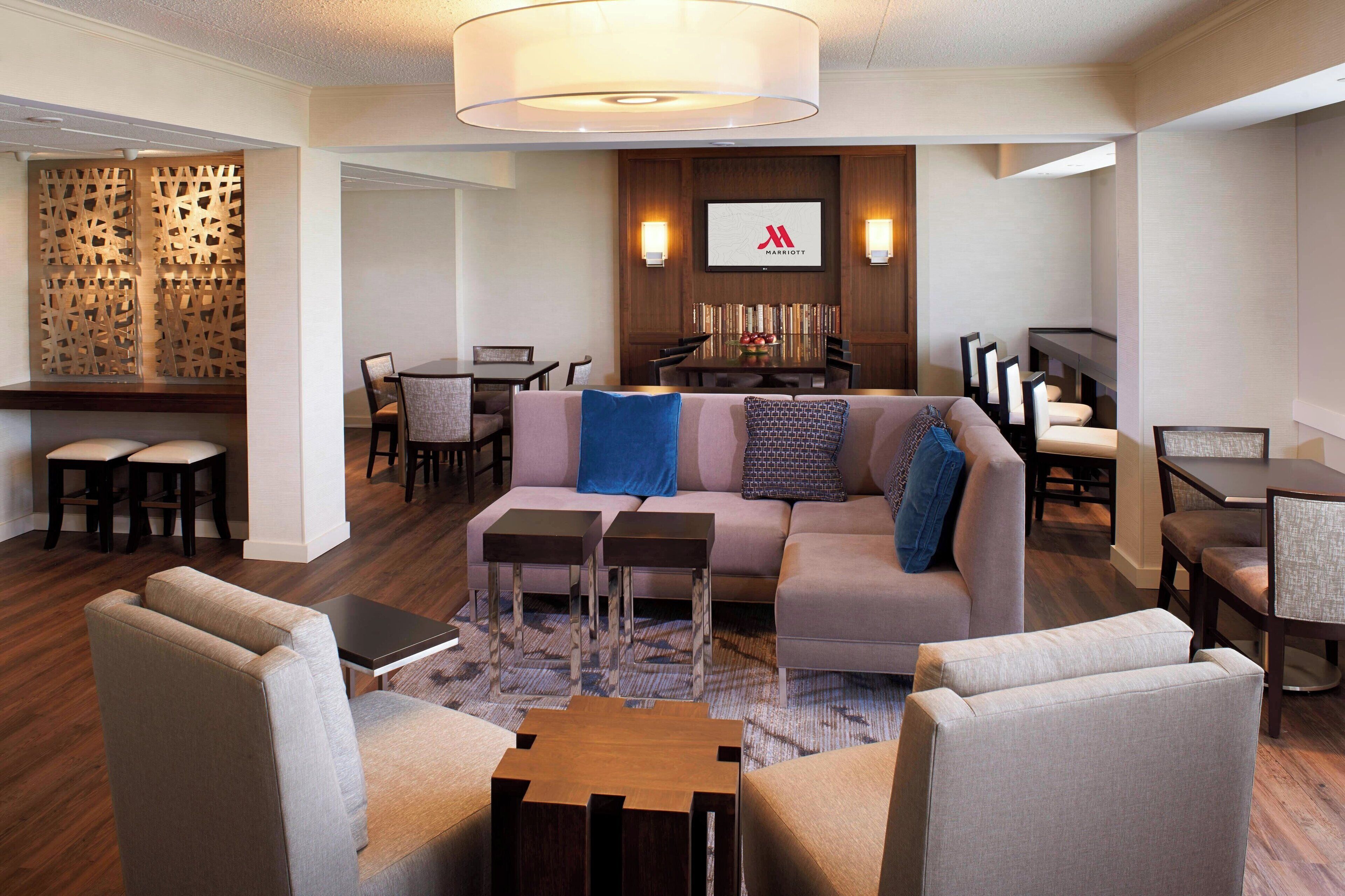 A modern hotel lobby with a sofa, chairs, a dining area, and a large screen displaying the Marriott logo.