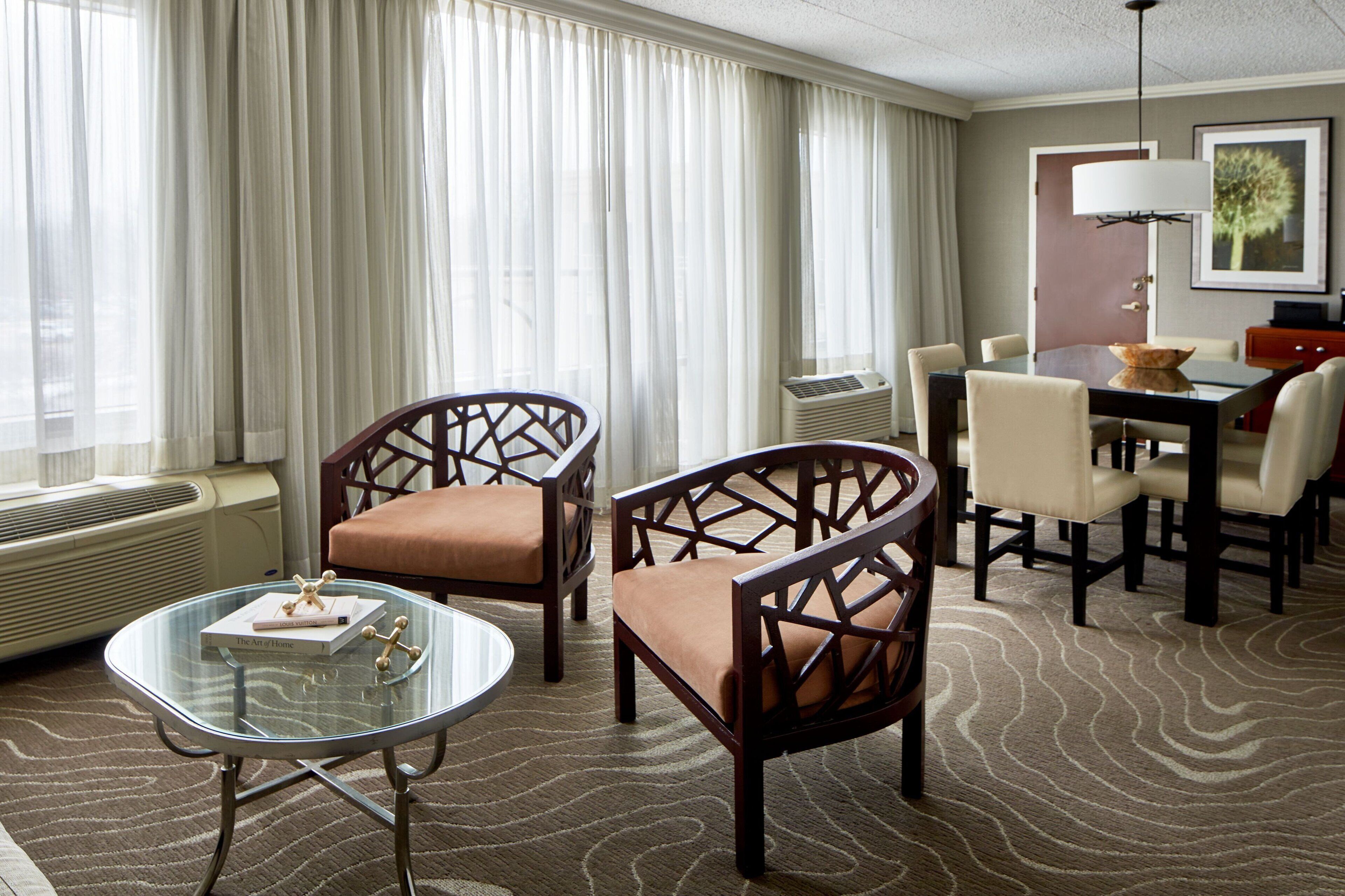 A hotel room with a dining area, a glass table, and two wooden chairs with cushions.