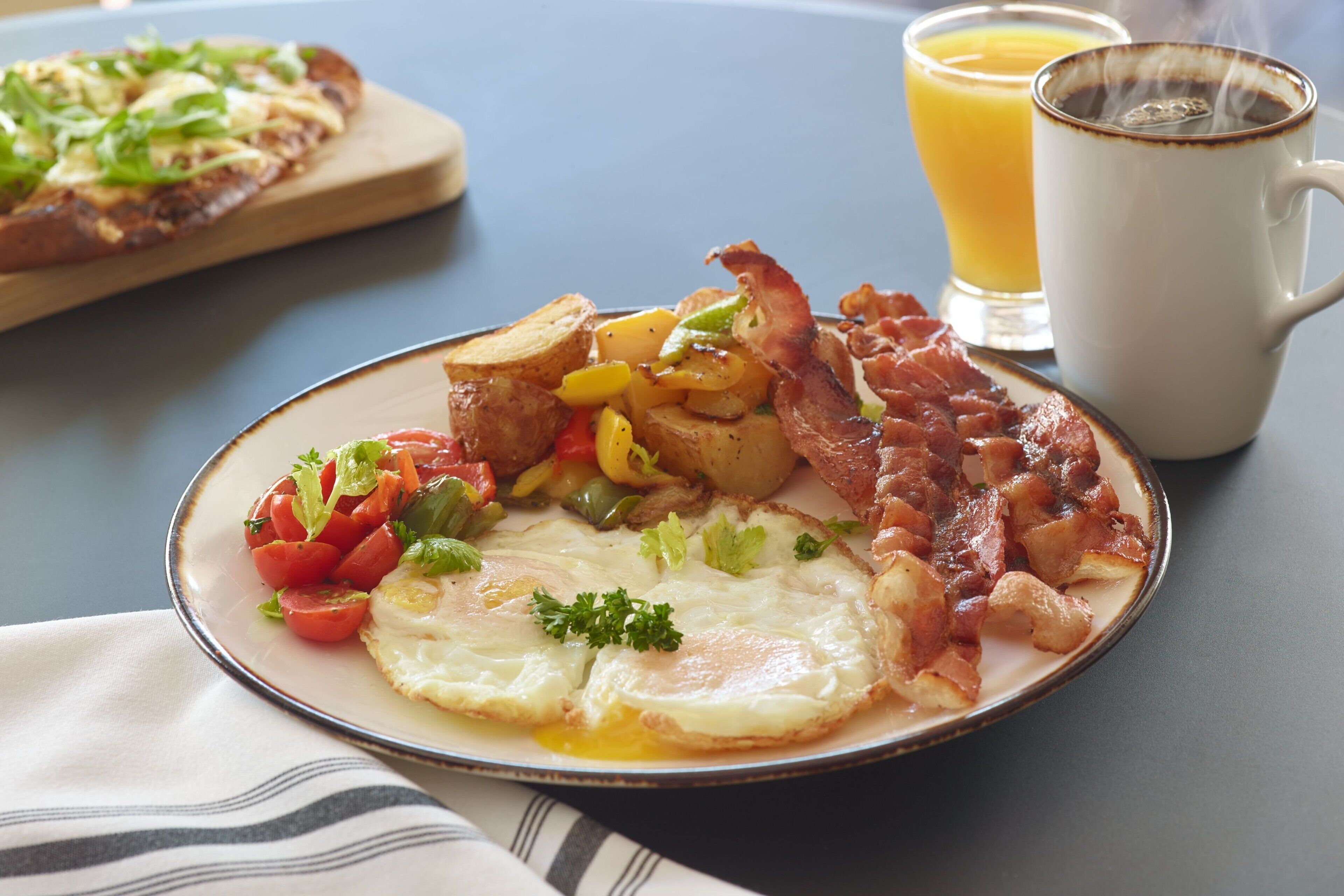 A breakfast plate with eggs, bacon, potatoes, and vegetables, a glass of orange juice, and a mug of coffee on a table.