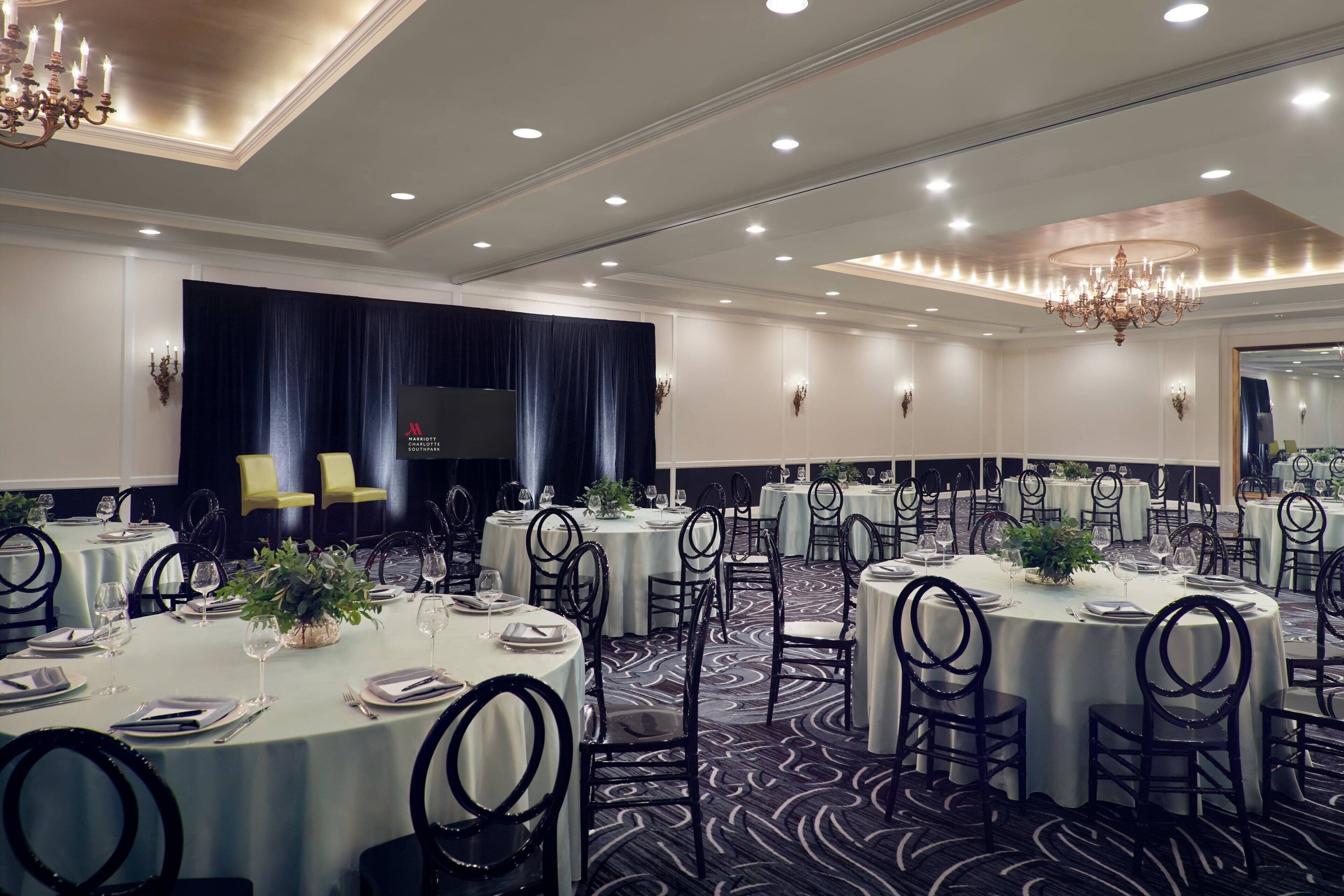 A banquet hall with round tables, black chairs, and chandeliers.