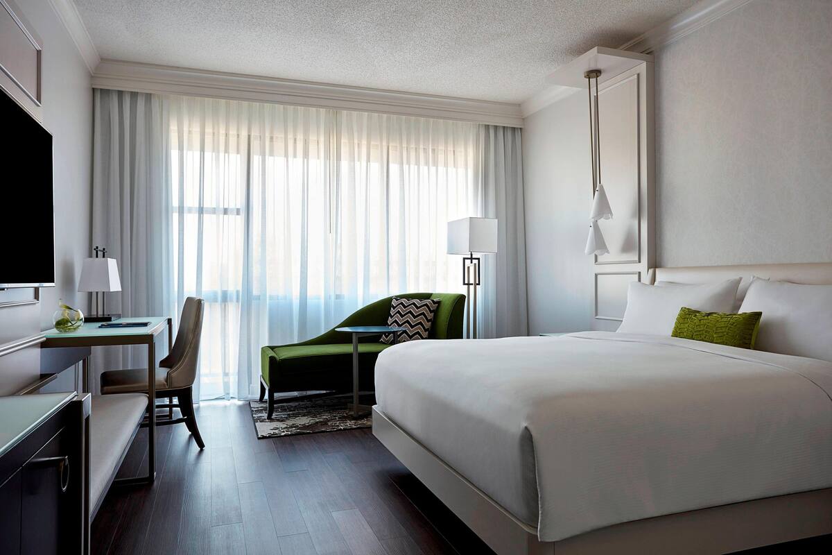 A hotel room with a large bed, a desk with a chair, a TV, and a window with sheer curtains.