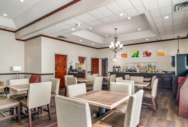 A hotel dining area with multiple tables and chairs, a bar, and a kitchen in the background.