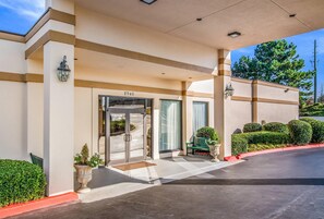 Entrada de hotel con puerta de cristal, el número '2945' sobre la puerta, un banco y plantas en macetas.