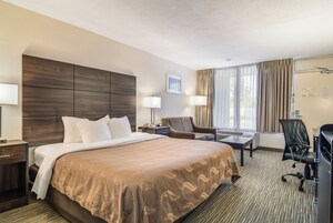 Habitación de hotel con una cama grande, dos mesitas de noche, un escritorio con una silla, una lámpara de escritorio, una silla adicional y un ventanal con cortinas.