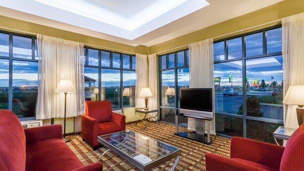 Habitación de hotel con un ventanal grande, un televisor de pantalla plana, sillas tapizadas en rojo, una mesa de centro de vidrio y una alfombra estampada.