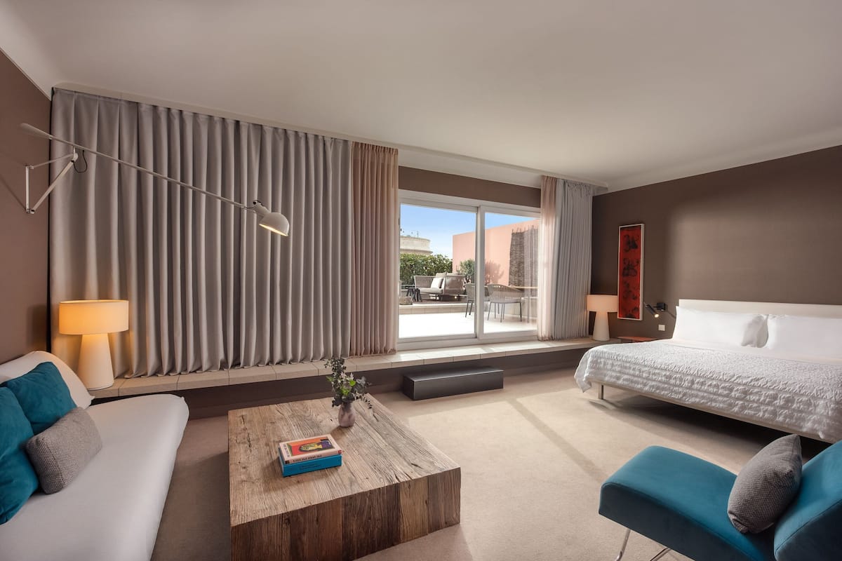 A modern hotel room with a bed, a sofa, a coffee table, and a large window with curtains.