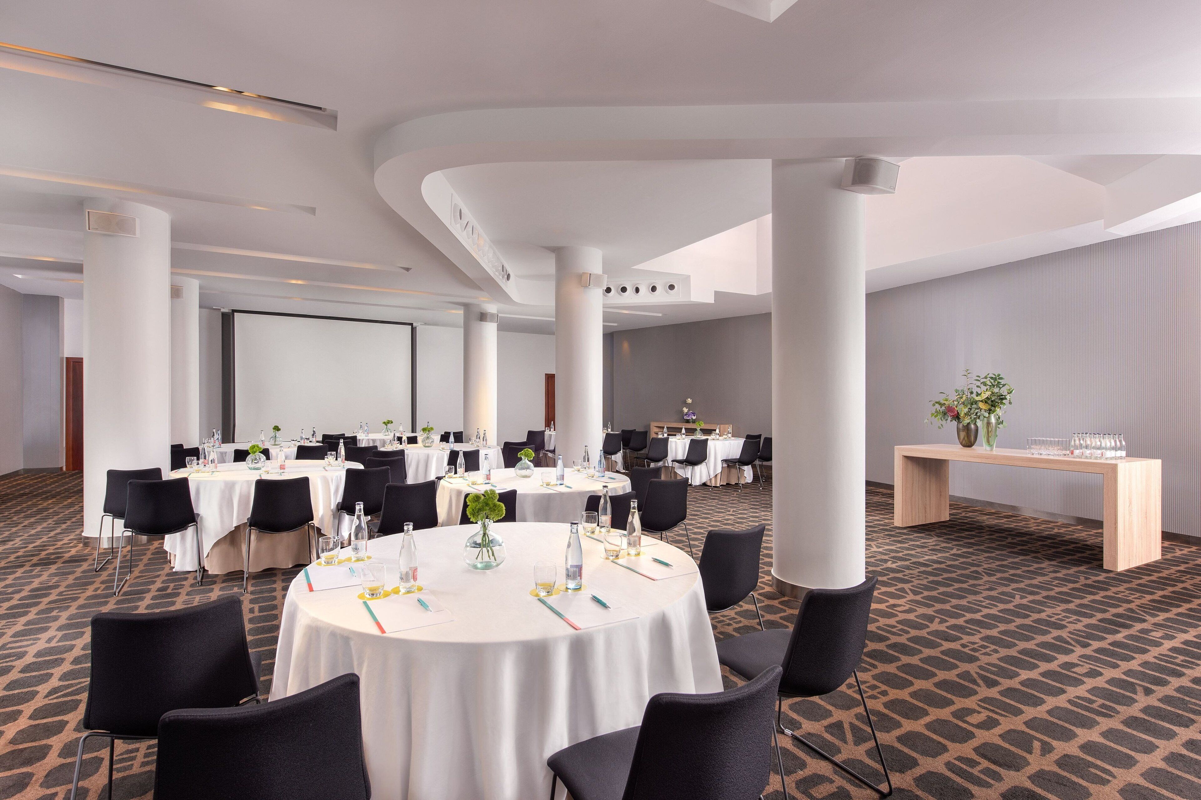 Un salón de conferencias con varias mesas redondas, sillas negras y una pantalla de proyector.