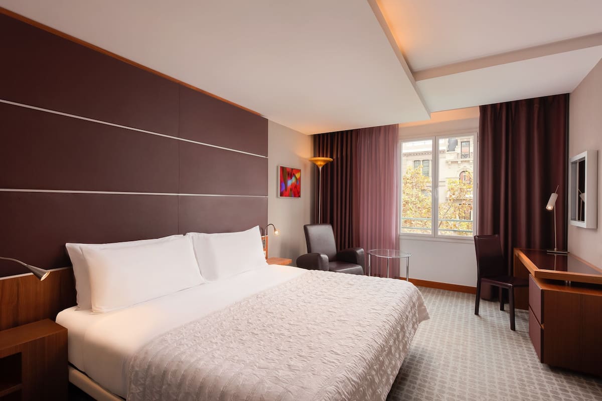A hotel room with a large bed, a desk, a chair, and a window with curtains.