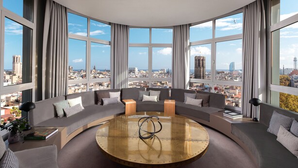 Una sala de estar moderna con una disposición de sofás curvos, una mesa de centro y ventanas amplias con vista a la ciudad.