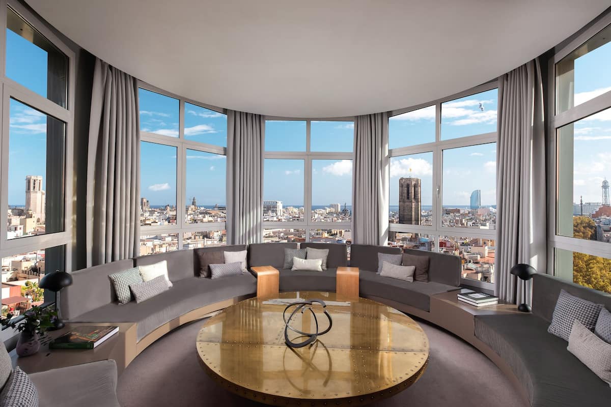 A modern living room with a curved sofa arrangement, a central coffee table, and large windows offering a city view.