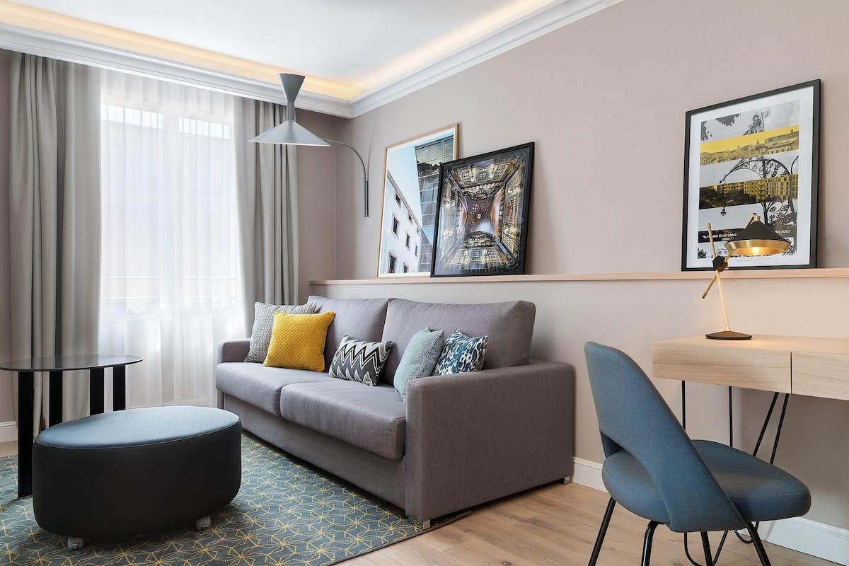A modern living room with a grey sofa, a round ottoman, a blue chair, a small table, and a desk with a lamp. There are two framed pictures on the wall.