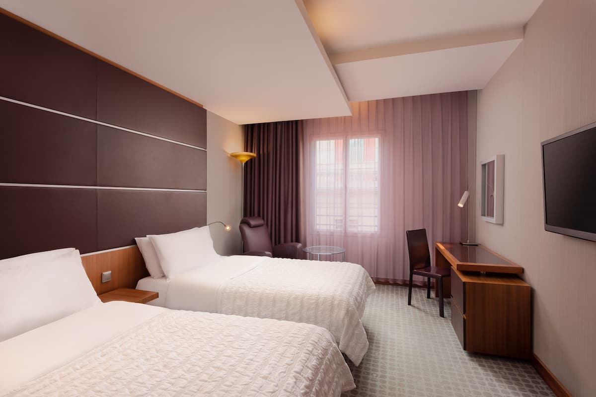 A hotel room with two beds, a desk, a chair, a TV, and a window with curtains.