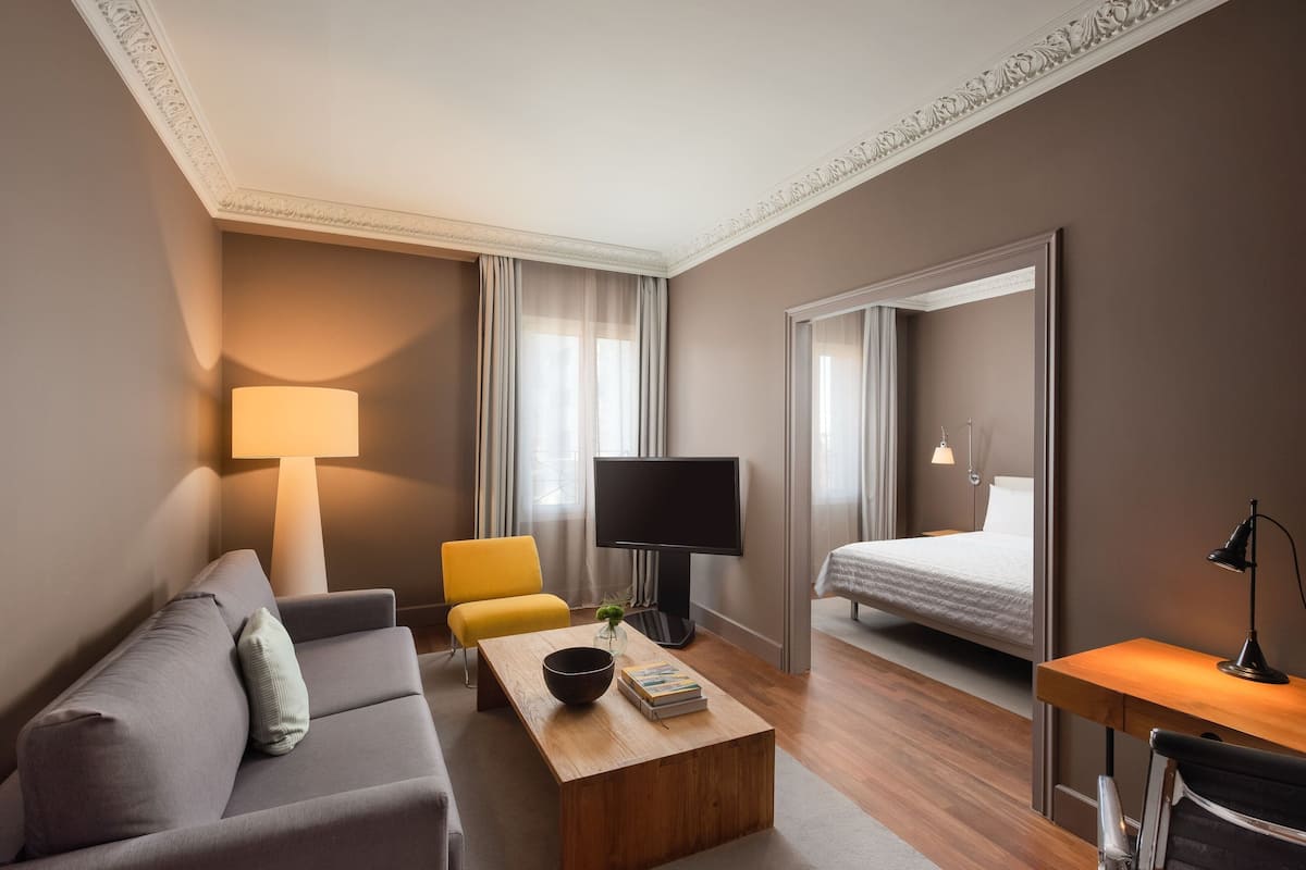 A modern hotel room with a sofa, a yellow chair, a wooden coffee table, a TV, a bed, and a desk with a lamp.