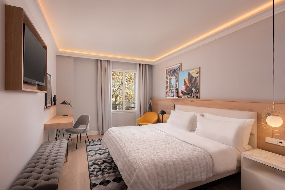 A modern hotel room with a large bed, a desk, a chair, and a bench.