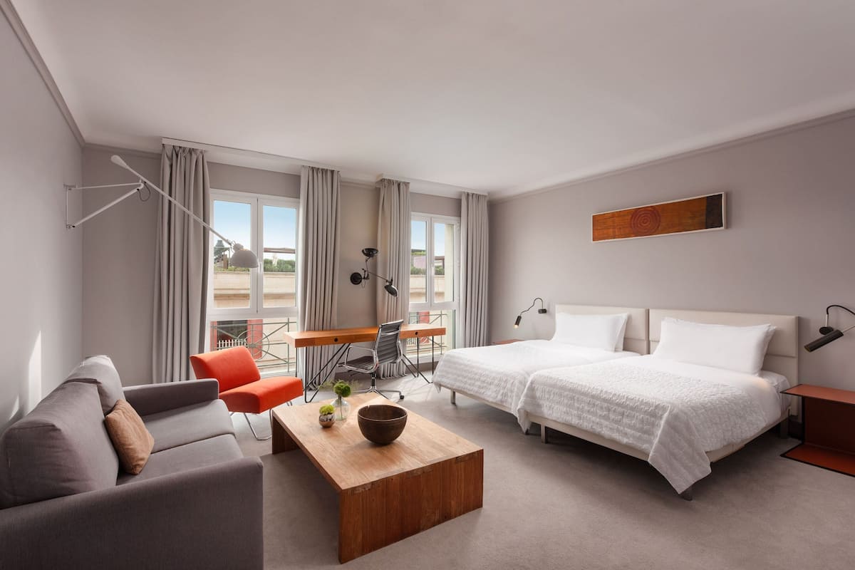 A modern hotel room with a sofa, two single beds, a desk with a chair, a small table with a bowl, and a view of the outdoors through large windows.