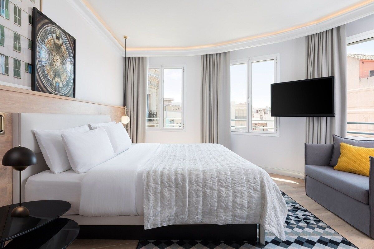 A modern hotel room with a large bed, a flat-screen TV, a sofa with a yellow pillow, and a large window offering a city view.