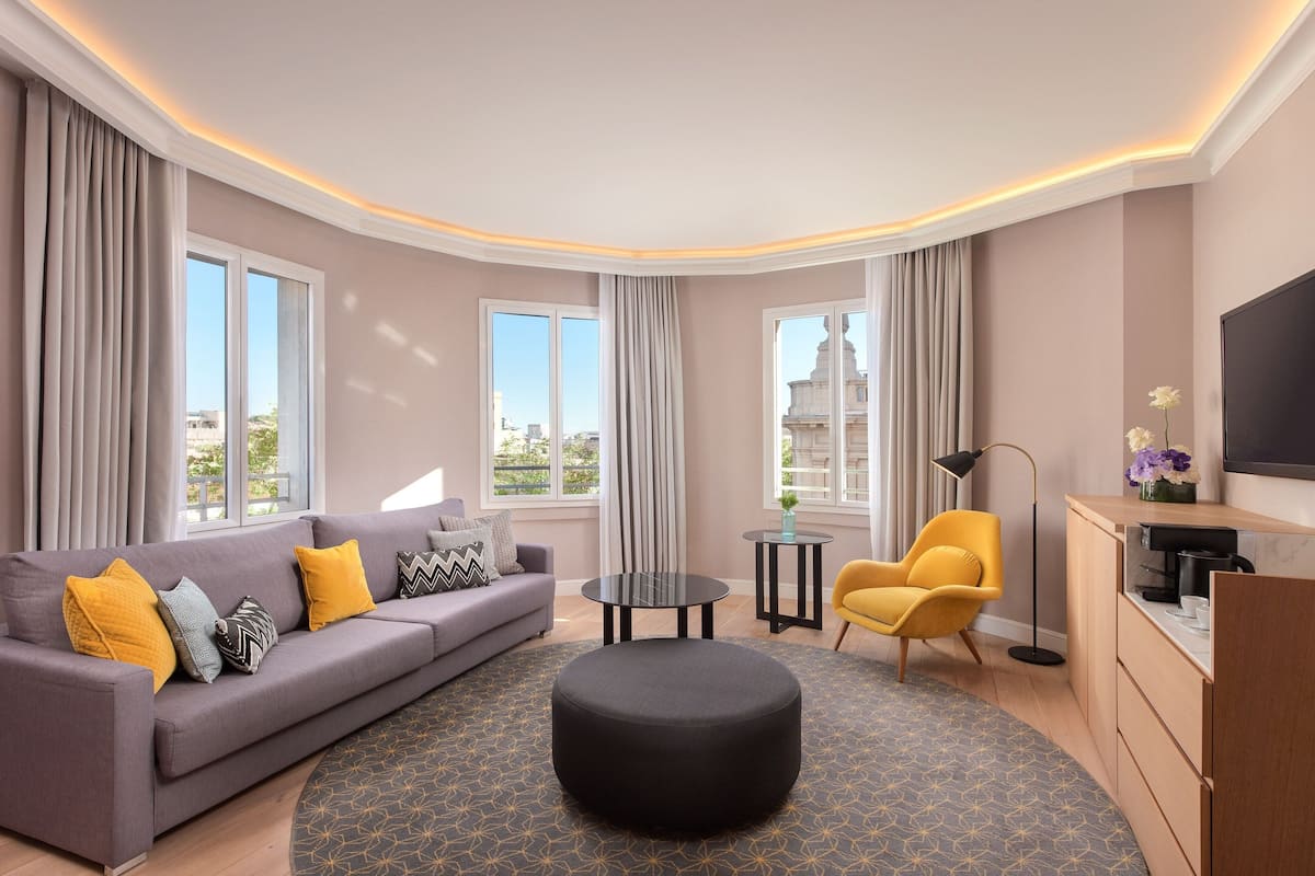 A modern living room with a grey sofa, yellow and grey cushions, a black ottoman, and a patterned rug. There are two windows with curtains, a small round table, and a yellow armchair.