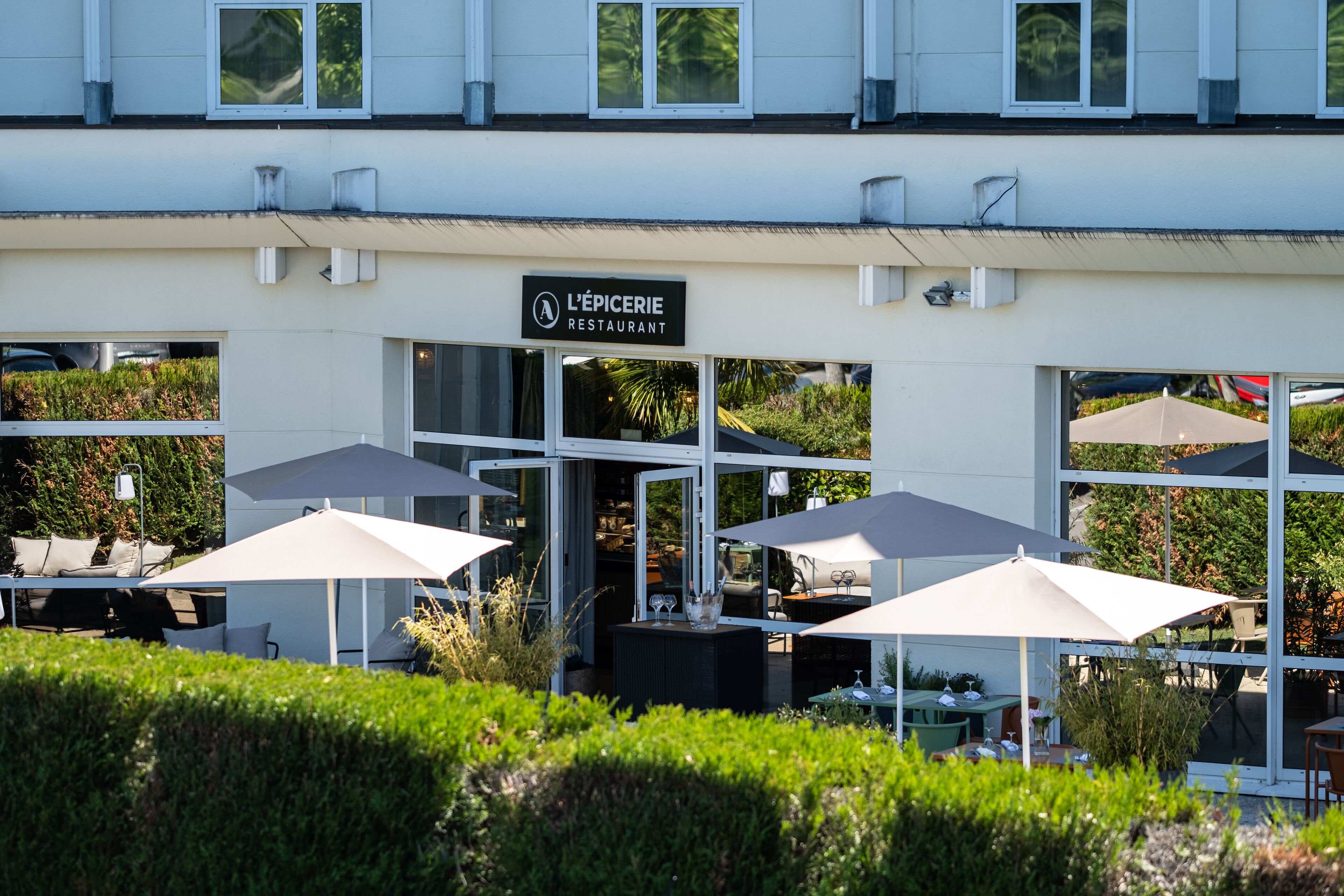 Un área de asientos al aire libre con sombrillas y mesas, un letrero que dice 'L'Épicerie Restaurant' y un edificio con ventanas grandes.